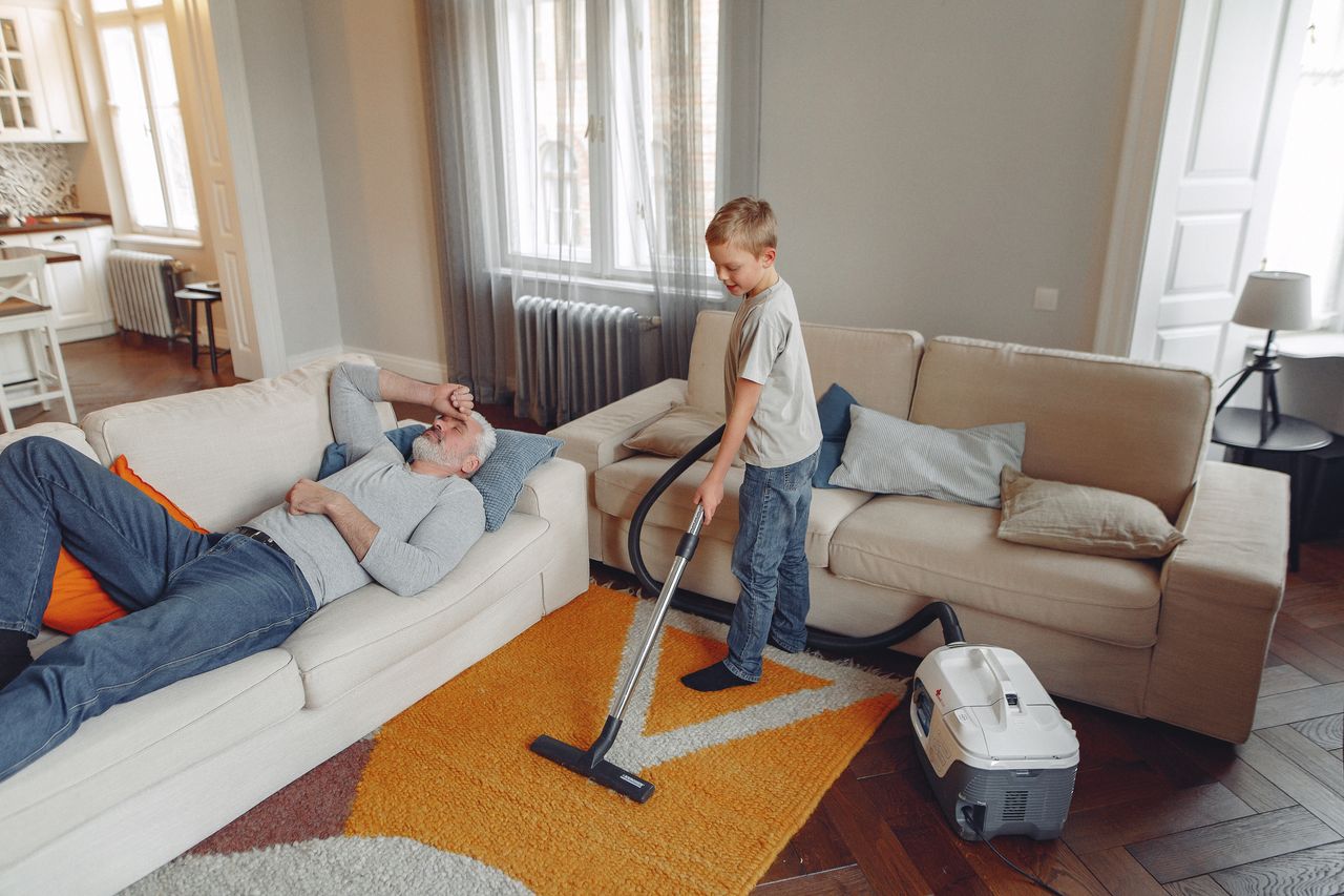 our-story A young boy is vacuuming the living room carpet while his grandfather rests on the sofa.